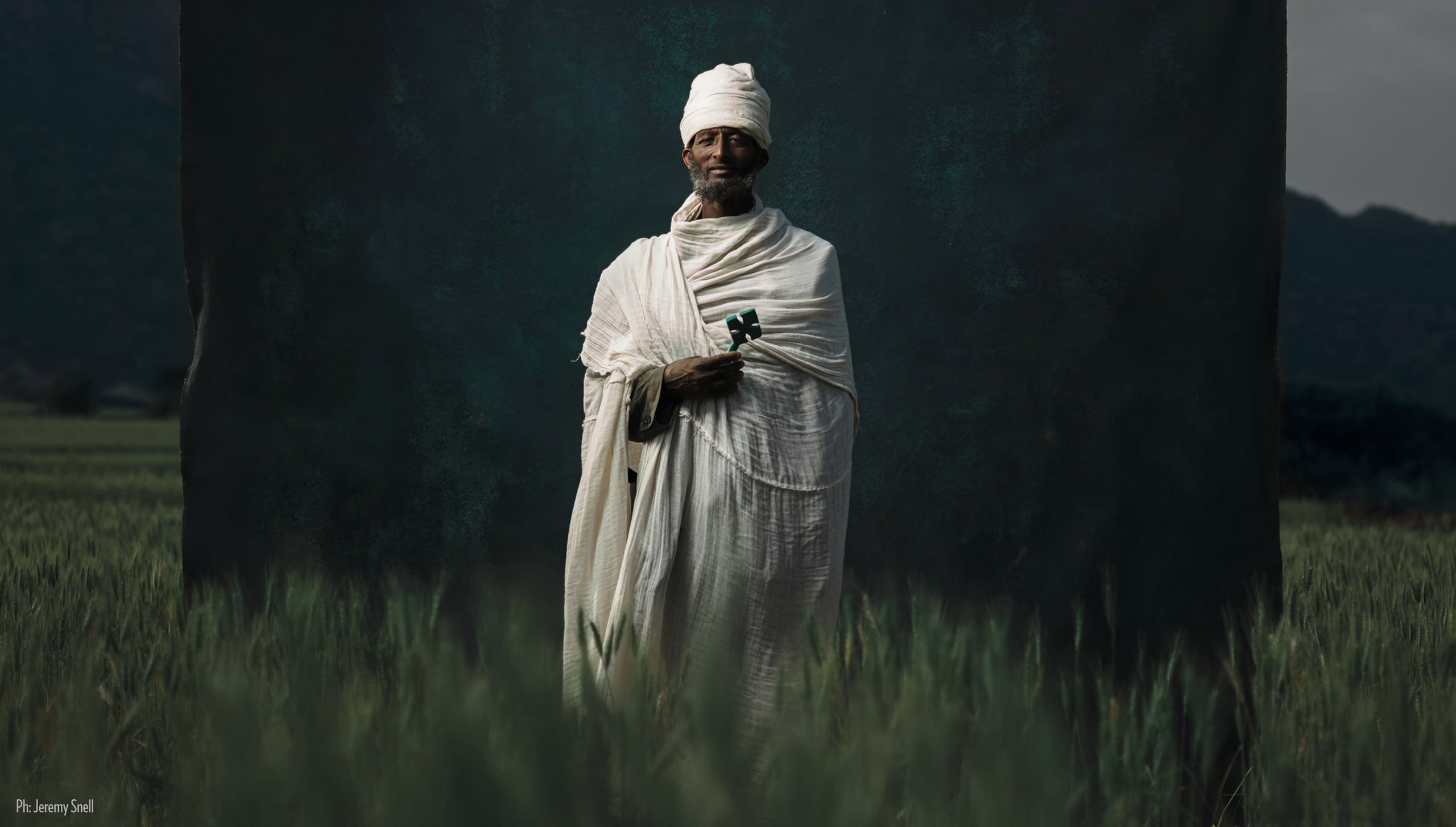 A man in a white robe stands in a field in front of a dark hand-painted canvas backdrop.